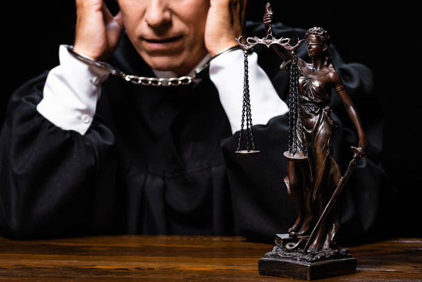 przycięty widok sędziego w szlafroku sądowym siedzącego przy stole z kajdankami odizolowanymi na czarno - Zdjęcie, obraz