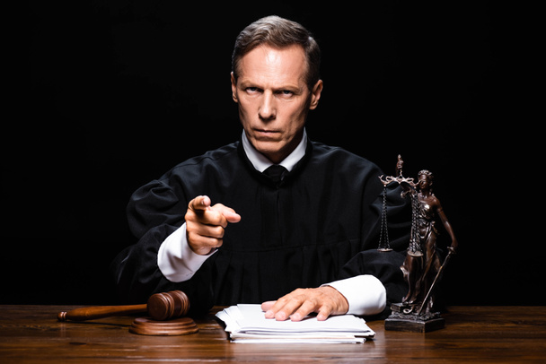 judge in judicial robe sitting at table and pointing with finger isolated on black - Photo, Image