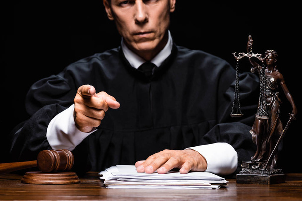 przycięty widok sędziego w szacie sądowej siedzącego przy stole i wskazującego palcem odizolowanym na czarno - Zdjęcie, obraz