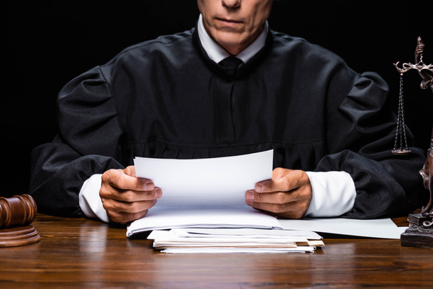 cropped view of judge in judicial robe sitting at table and reading paper isolated on black - Фото, зображення