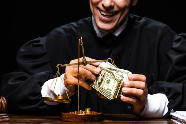 przycięty widok uśmiechniętego sędziego w szlafroku sądowym siedzącego przy stole i trzymającego banknoty dolarowe odizolowane na czarno - Zdjęcie, obraz