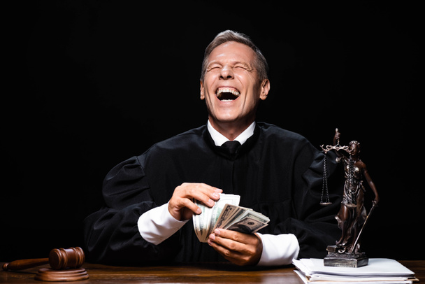 uśmiechnięty sędzia w szlafroku sądowym siedzący przy stole i trzymający banknoty dolarowe odizolowane na czarno - Zdjęcie, obraz