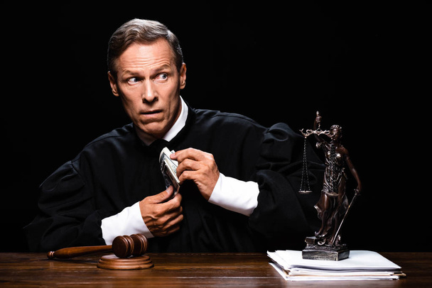 Yargı bornozu giymiş ürkek bir yargıç masada oturuyor ve elinde siyah banknotlar tutuyor. - Fotoğraf, Görsel