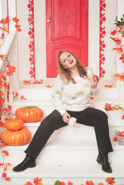 Bastante joven mujer de tamaño grande en suéter blanco acogedor bebiendo el primer café del día sentado en el porche de una hermosa casa de otoño con una puerta roja
 - Foto, imagen