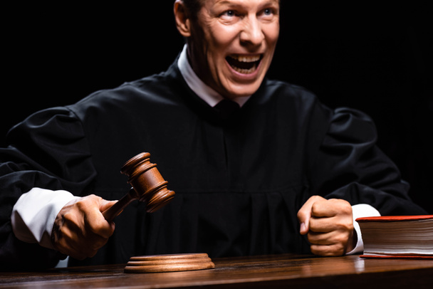wściekły sędzia w szacie sądowej siedzi przy stole i uderza młotkiem odizolowanym na czarno - Zdjęcie, obraz