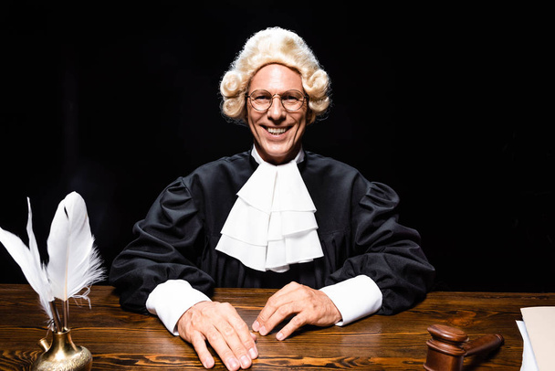 sonriente juez en bata judicial y peluca sentado a la mesa aislado en negro
 - Foto, Imagen