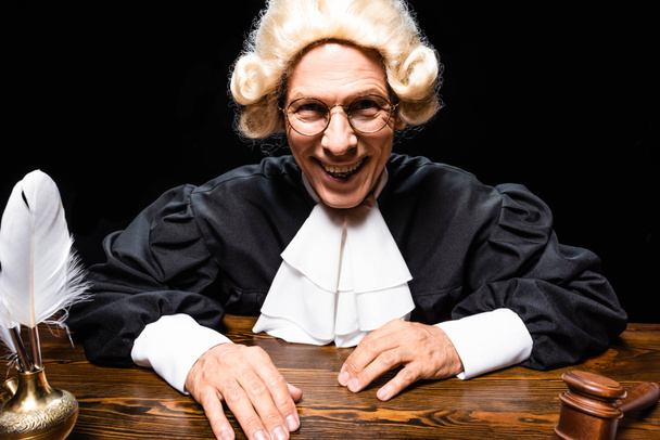 smiling judge in judicial robe and wig sitting at table isolated on black - 写真・画像