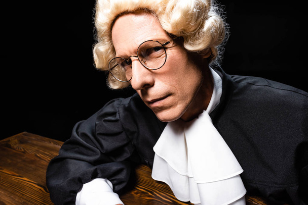 judge in judicial robe and wig sitting at table and looking at camera isolated on black - 写真・画像
