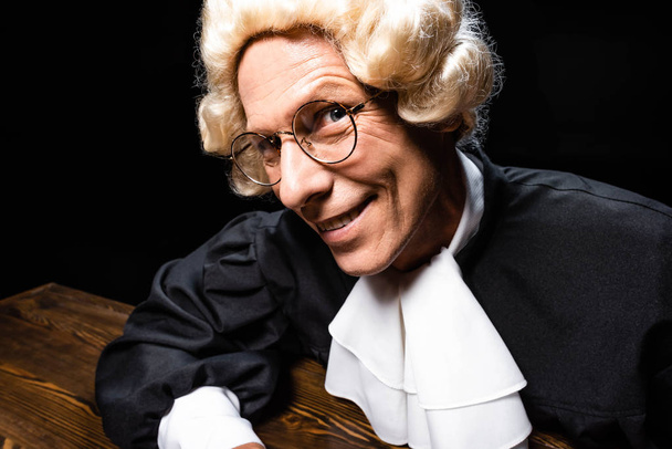 giudice sorridente in veste giudiziaria e parrucca seduta a tavola isolata su nero
 - Foto, immagini