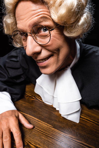 mosolygó bíró a bírósági köntös és paróka ül az asztalnál elszigetelt fekete - Fotó, kép