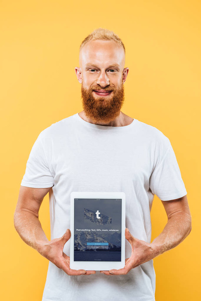 Kiev, Oekraïne - 27 augustus 2019: lachende man met baard toont digitale tablet met tuimelaar app op scherm, geïsoleerd op geel - Foto, afbeelding