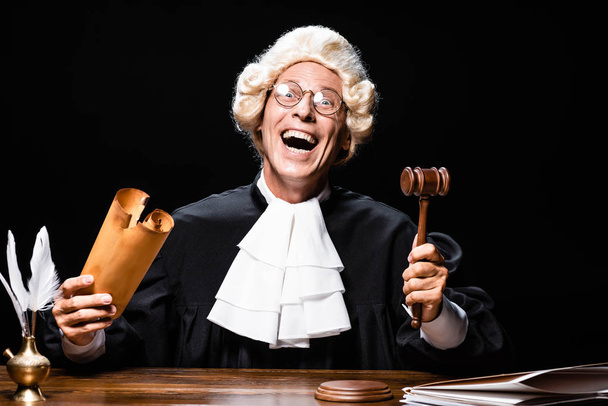 uśmiechnięty sędzia w szlafroku sądowym i peruce siedzący przy stole, trzymający dokument i młotek odizolowany na czarno - Zdjęcie, obraz