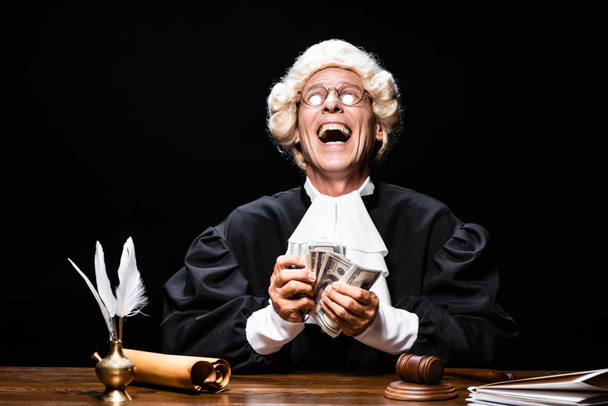 χαμογελαστός δικαστής με δικαστική ρόμπα και περούκα κάθεται στο τραπέζι και κρατά τα χρήματα που απομονώνονται σε μαύρο - Φωτογραφία, εικόνα