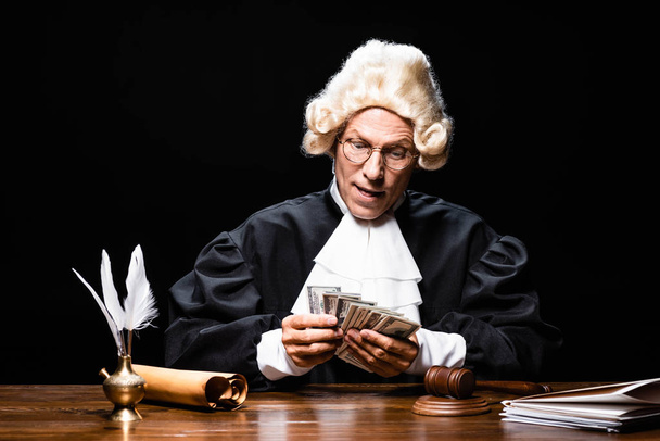 rechter in gerechtelijk gewaad en pruik zitten aan tafel en tellen geld geïsoleerd op zwart - Foto, afbeelding