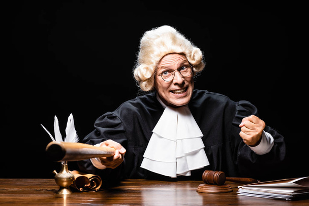 angry judge in judicial robe and wig sitting at table and holding bat isolated on black - Photo, Image