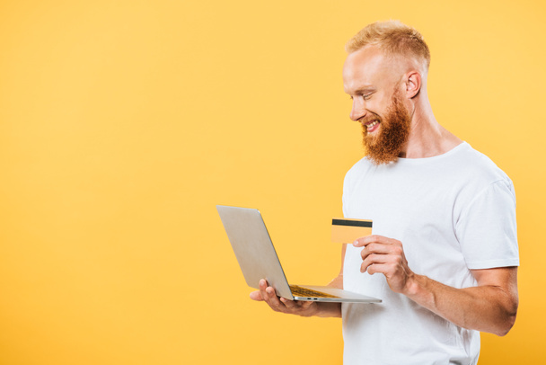 cheerful bearded man shopping online with laptop and credit card, isolated on yellow - Φωτογραφία, εικόνα