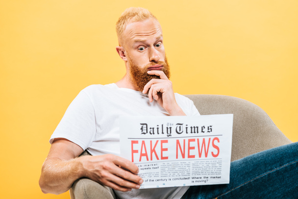 thoughtful man reading newspaper with fake news while sitting on armchair, isolated on yellow - Foto, Imagem