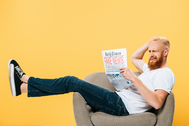 frustrated bearded man reading newspaper with fake news while sitting on armchair, isolated on yellow - Foto, imagen