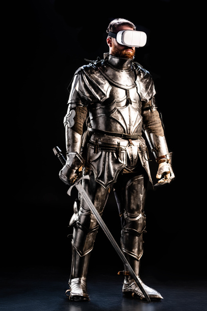 knight with virtual reality headset in armor holding sword isolated on black  - Photo, Image