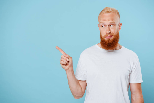 portrait of sarcastic bearded man in white t-shirt pointing at something, isolated on blue  - Fotografie, Obrázek