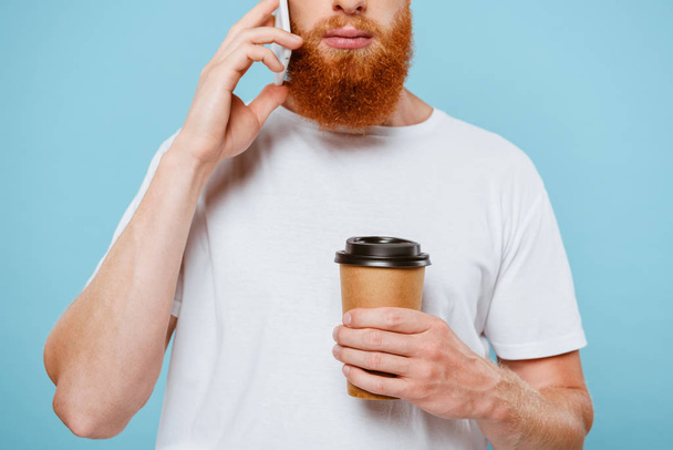 cropped view of serious bearded man with coffee to go talking on smartphone, isolated on blue - Photo, Image