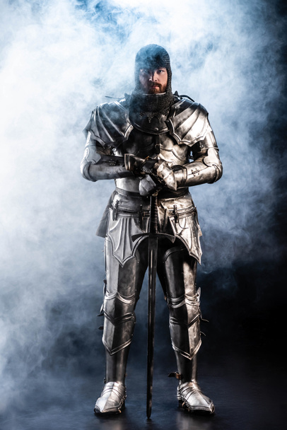 handsome knight in armor looking at camera and holding sword on black background  - Фото, зображення