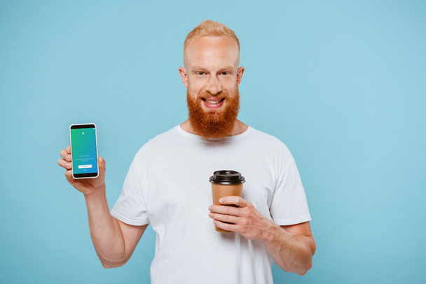 KYIV, UKRAINE - AUGUST 27, 2019: smiling bearded man with coffee to go showing smartphone with twitter app, isolated on blue - Photo, Image