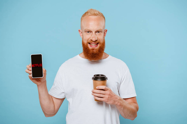 KYIV, UKRAINE - AUGUST 27, 2019: smiling bearded man with coffee to go showing smartphone with netflix app, isolated on blue - Fotografie, Obrázek