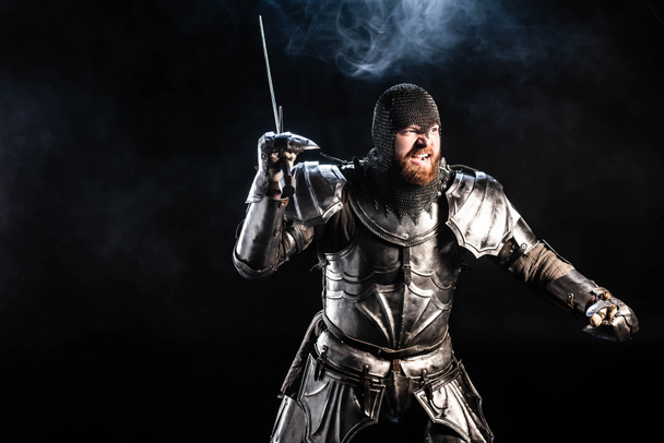 Siyah arka planda kılıçla savaşan zırhlı yakışıklı ve kızgın bir şövalye.  - Fotoğraf, Görsel
