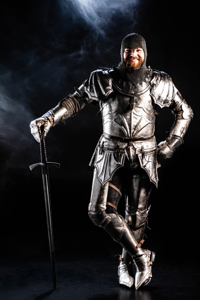sonriente caballero con armadura mirando a la cámara y sosteniendo la espada sobre fondo negro
  - Foto, Imagen