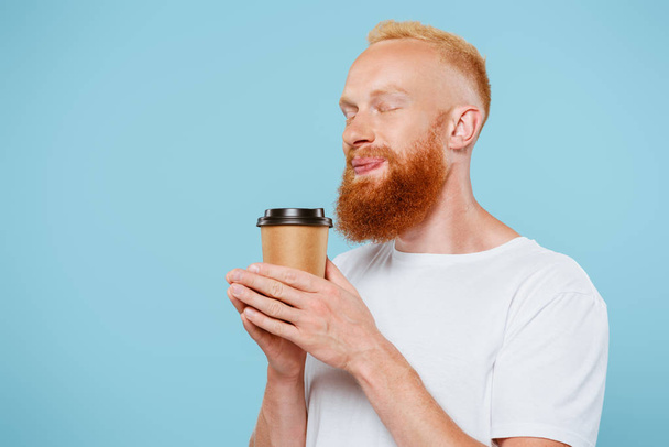 smiling bearded man in t-shirt holding coffee to go with closed eyes, isolated on blue - Foto, Imagen