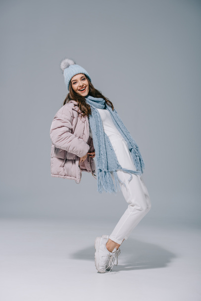 attractive smiling girl dancing in warm coat, knitted hat and scarf on grey - Foto, afbeelding