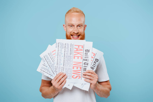 happy bearded man holding newspapers with fake news, isolated on blue - Photo, image
