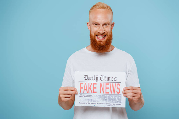 smiling bearded man showing newspaper with fake news, isolated on blue - Fotoğraf, Görsel