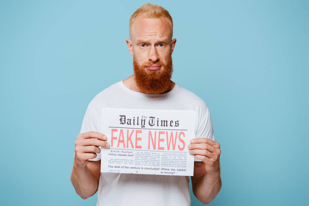 upset bearded man showing newspaper with fake news, isolated on blue - Foto, Imagen