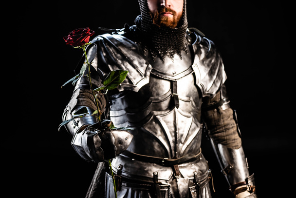 Обрезанный вид рыцаря в доспехах, держащего розу, изолированный на черном
  - Фото, изображение