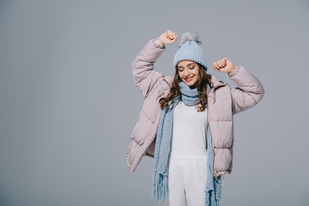 привлекательная счастливая женщина, танцующая в теплом пальто, вязаной шляпе и шарфе, изолированная на сером
 - Фото, изображение