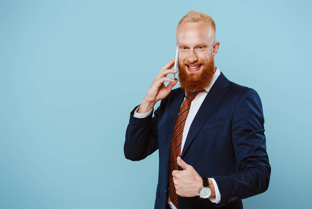 smiling bearded businessman talking on smartphone and showing thumb up, isolated on blue - Foto, immagini