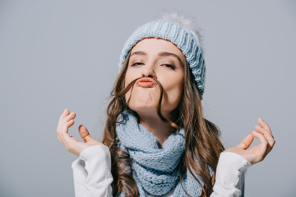 beautiful funny girl in knitted hat and scarf making mustache from hair, isolated on grey - Photo, Image