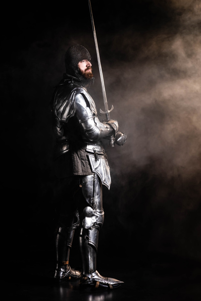 handsome knight in armor holding sword on black background - Foto, afbeelding