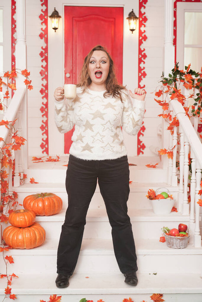 Bastante joven mujer de tamaño grande en suéter blanco acogedor bebiendo el primer café del día sentado en el porche de una hermosa casa de otoño con una puerta roja
 - Foto, imagen