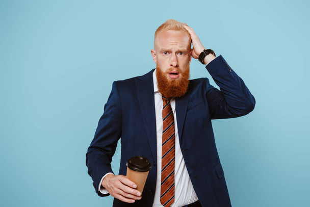 worried bearded businessman holding coffee to go, isolated on blue - Фото, зображення
