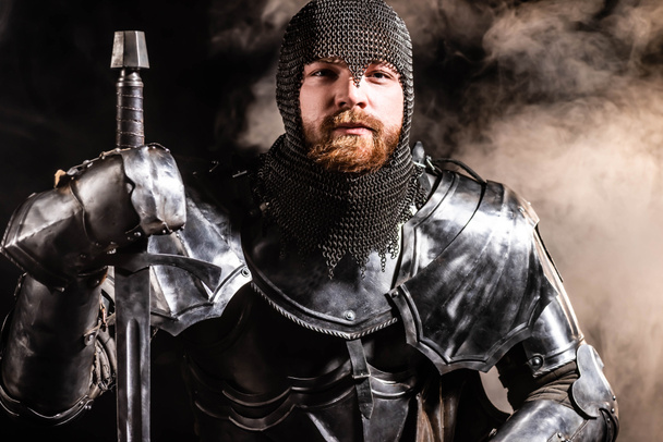 handsome knight in armor holding sword on black background - Photo, Image