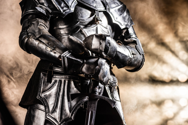 cropped view of knight in armor holding sword on black background - Fotó, kép