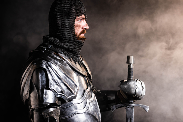 zijaanzicht van knappe ridder in harnas met zwaard op zwarte achtergrond - Foto, afbeelding