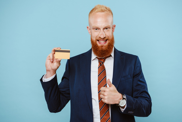 handsome cheerful businessman holding credit card and showing thumb up, isolated on blue - Foto, Imagem