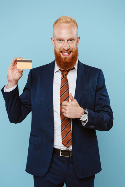 bearded smiling businessman holding credit card and showing thumb up, isolated on blue - Foto, afbeelding