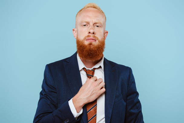 confident bearded businessman in suit correcting tie, isolated on blue - Foto, imagen