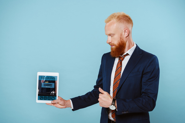 bearded businessman in suit showing digital tablet with booking app, isolated on blue - Foto, afbeelding