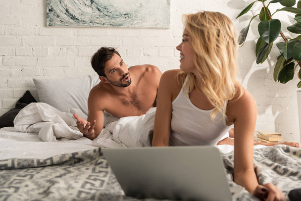attractive smiling woman using laptop in bed with boyfriend quarreling behind - Fotó, kép